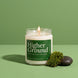 Higher Ground 8 oz soy candle by Upsensed, lit, in a clear glass jar with a green label, beside a tuft of moss and a wet black stone with water droplets on a green backdrop.