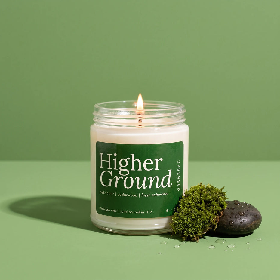 Higher Ground 8 oz soy candle by Upsensed, lit, in a clear glass jar with a green label, beside a tuft of moss and a wet black stone with water droplets on a green backdrop.