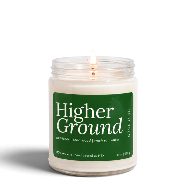 Higher Ground 8 oz soy candle by Upsensed, lit, in a clear glass jar with a forest green label listing petrichor, cedarwood, and fresh rainwater notes.