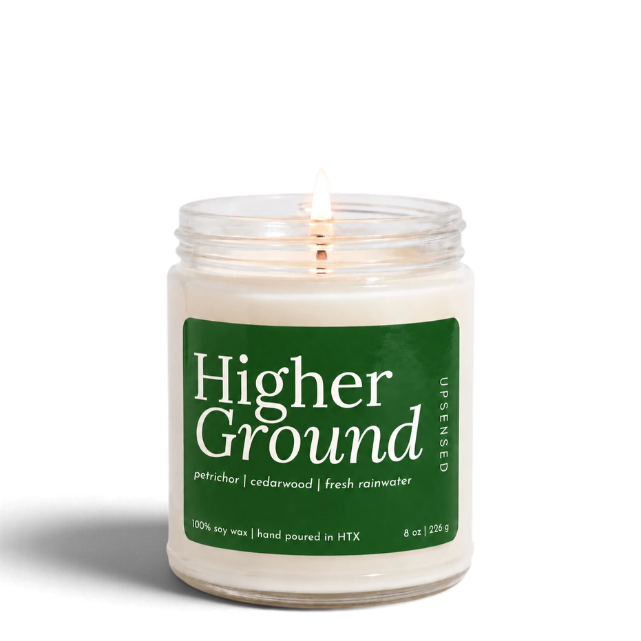 Higher Ground 8 oz soy candle by Upsensed, lit, in a clear glass jar with a forest green label listing petrichor, cedarwood, and fresh rainwater notes.