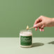 A hand with gold rings and pink nails lighting a Higher Ground 8 oz soy candle by Upsensed with a wooden match, on a two-tone green set with soft sunlight.
