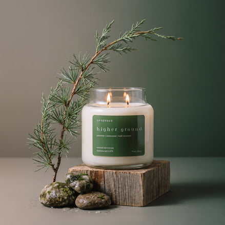 Upsensed Higher Ground candle burning on a weathered wood block, styled with a juniper branch and wet, moss-covered stones against a green background.