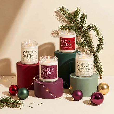 Holiday candle gift set of four lit 8 oz soy candles, Shortbread + Sage, Berry Bright, Fir + Cider and Velvet Spruce, on red, plum, green and sage plinths with a spruce branch, string lights and glass ornaments
