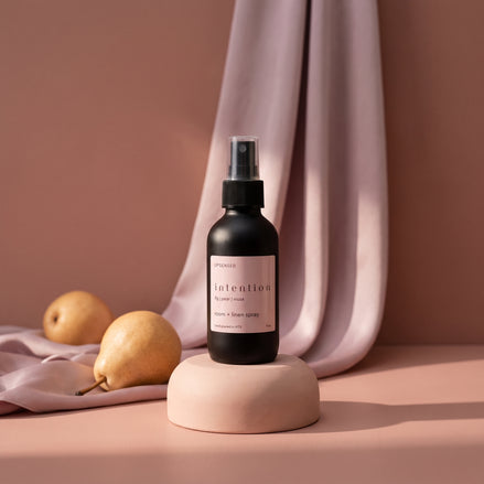 Intention room and linen spray on a blush pedestal with draped mauve satin and two pears.