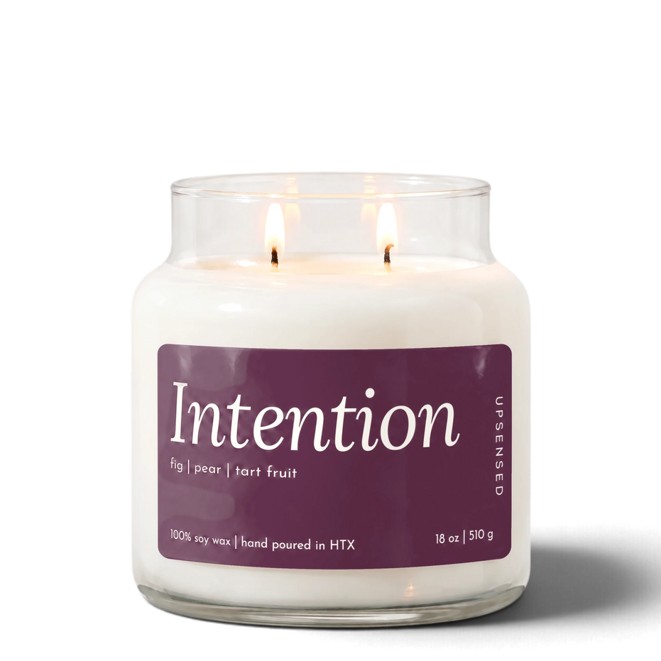 Intention 18 oz double-wick soy candle by Upsensed, both wicks lit, in a clear glass jar with a pink label listing fig, pear, and tart fruit notes.
