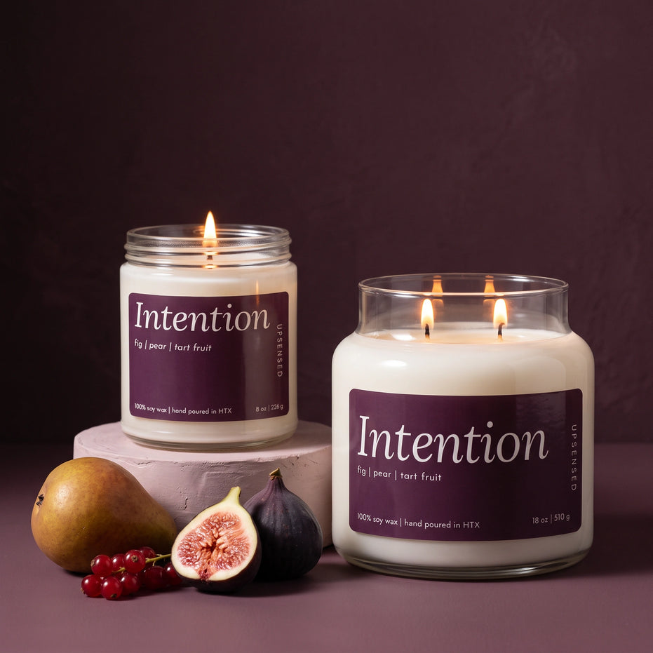 Intention 8 oz and 18 oz soy candles by Upsensed, lit, with deep fig purple labels, the 8 oz on a blush plaster riser beside a ripe pear, a halved fig, a whole fig, and red currants against a dark plum backdrop.