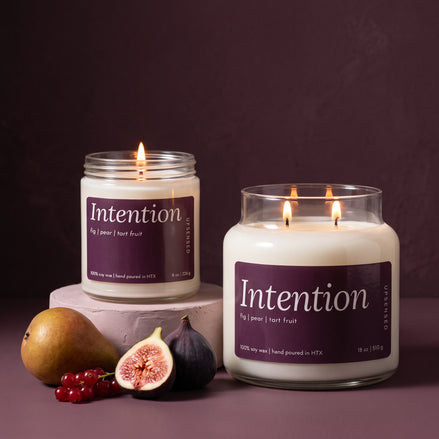 Intention 8 oz and 18 oz soy candles by Upsensed, lit, with deep fig purple labels, the 8 oz on a blush plaster riser beside a ripe pear, a halved fig, a whole fig, and red currants against a dark plum backdrop.