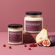 Intention soy candles by Upsensed in 8 oz and 18 oz sizes with black lids and plum labels listing fig, pear, and tart fruit notes, the 18 oz raised on a rose block, styled with a halved pear, halved figs, and red currants on a dusty rose set.