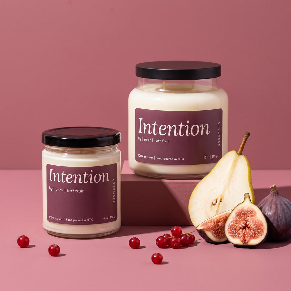 Intention soy candles by Upsensed in 8 oz and 18 oz sizes with black lids and plum labels listing fig, pear, and tart fruit notes, the 18 oz raised on a rose block, styled with a halved pear, halved figs, and red currants on a dusty rose set.