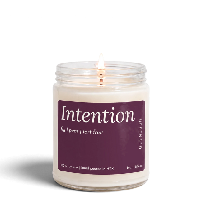 Intention 8 oz soy candle by Upsensed, lit, in a clear glass jar with a pink label listing fig, pear, and tart fruit notes.