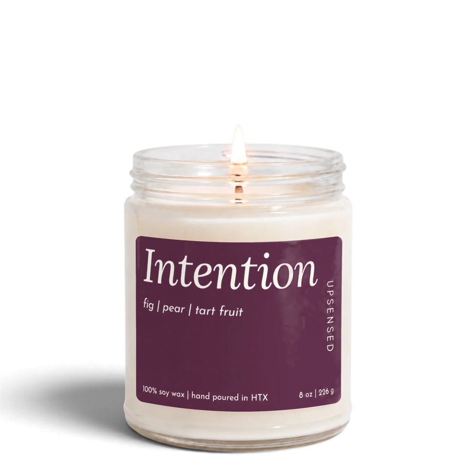 Intention 8 oz soy candle by Upsensed, lit, in a clear glass jar with a pink label listing fig, pear, and tart fruit notes.