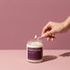 A hand with gold rings and blush pink nails lighting an Intention 8 oz soy candle by Upsensed with a wooden match, on a two-tone dusty rose set with soft sunlight.