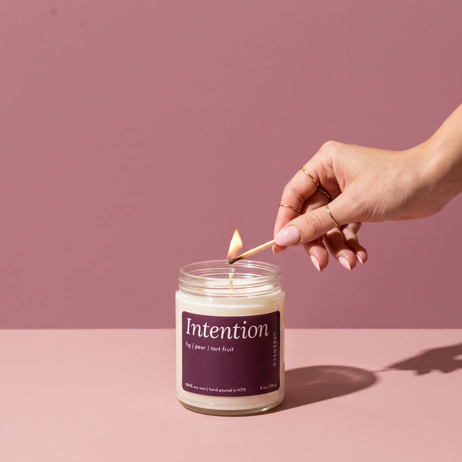 A hand with gold rings and blush pink nails lighting an Intention 8 oz soy candle by Upsensed with a wooden match, on a two-tone dusty rose set with soft sunlight.
