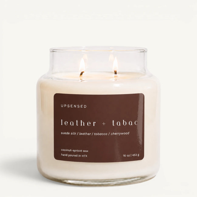 Upsensed Leather + Tabac 16 oz double-wick candle in a clear apothecary jar with a brown label, suede silk, leather, tobacco, and cherrywood scent notes, on a plain white background.