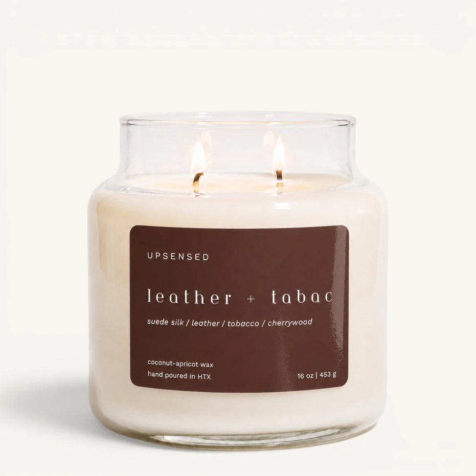 Upsensed Leather + Tabac 16 oz double-wick candle in a clear apothecary jar with a brown label, suede silk, leather, tobacco, and cherrywood scent notes, on a plain white background.