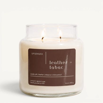 Upsensed Leather + Tabac 16 oz double-wick candle in a clear apothecary jar with a dark brown label, suede silk, leather, tobacco, and cherrywood scent notes, on a plain white background.