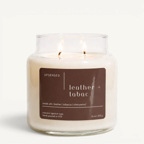 Upsensed Leather + Tabac 16 oz double-wick candle in a clear apothecary jar with a dark brown label, suede silk, leather, tobacco, and cherrywood scent notes, on a plain white background.