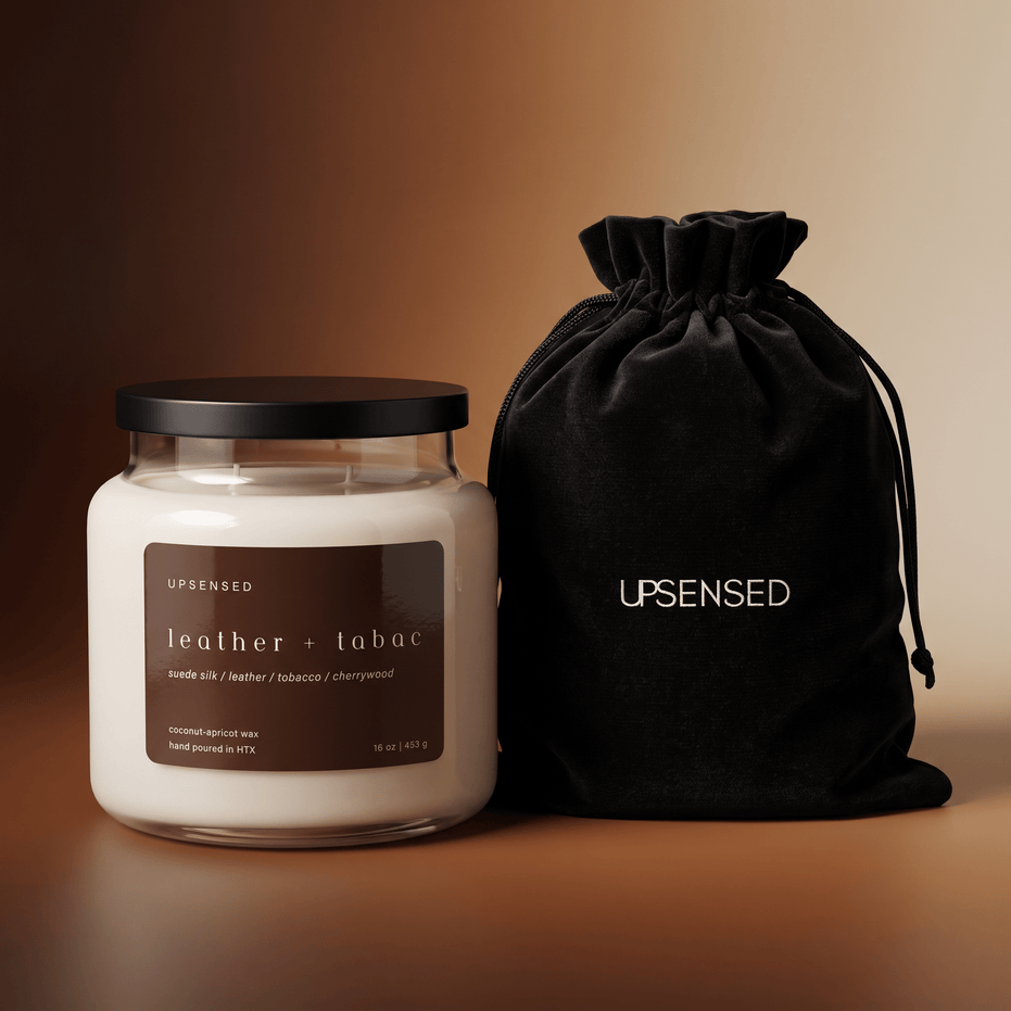 Upsensed Leather + Tabac candle with a black lid beside its black velvet UPSENSED drawstring packaging bag, on a warm brown gradient background.