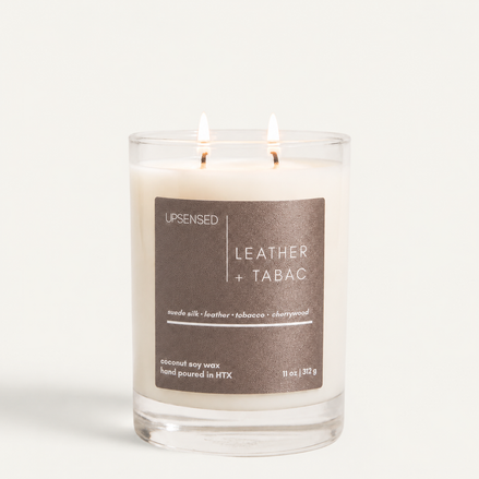 Leather + Tabac candle with dark brown label in a clear glass jar, two wicks lit.