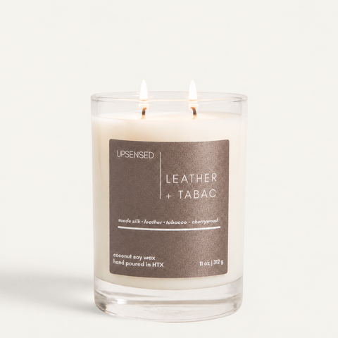 Leather + Tabac candle with dark brown label in a clear glass jar, two wicks lit.