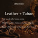 Leather + Tabac soy candle fragrance notes: suede silk, honey, and anise on top, leather, tobacco leaves, and cognac in the middle, and white musk, tonka bean, and cherrywood at the base, shown with brown leather, silk, and dried tobacco leaves on a dark wood table.