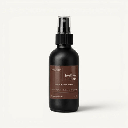Upsensed Leather + Tabac room and linen spray in a matte black 4 oz bottle with a brown label, suede silk, leather, tobacco, and cherrywood scent notes, on a plain white background.