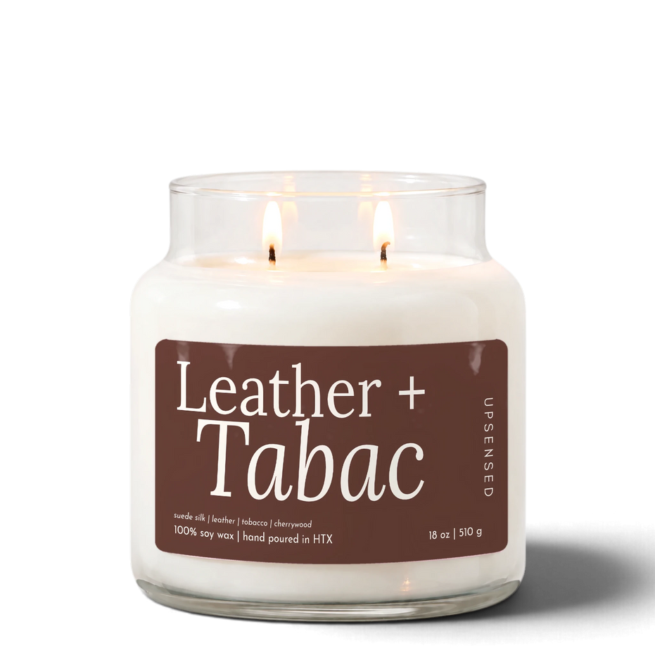 Leather + Tabac 18 oz double-wick soy candle by Upsensed, both wicks lit, in a clear glass jar with a brown label listing suede silk, leather, tobacco, and cherrywood notes.