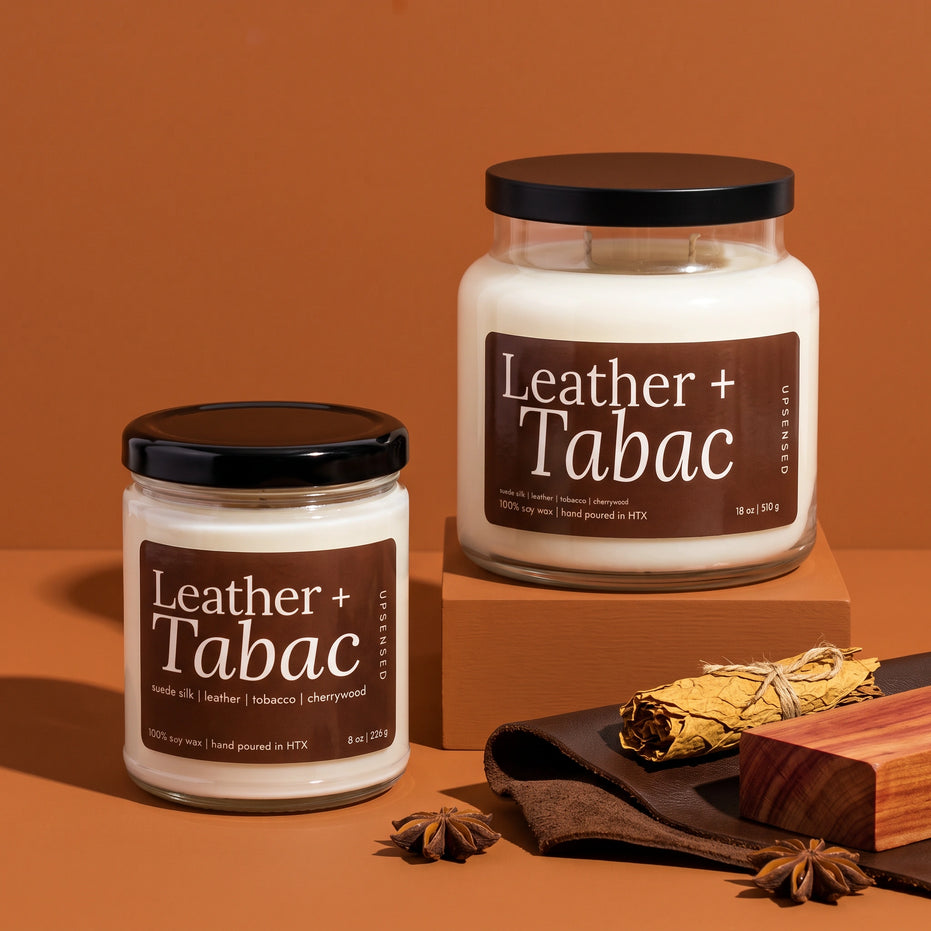 Leather + Tabac soy candles by Upsensed in 8 oz and 18 oz sizes with black lids and brown labels, the 18 oz raised on a cognac block, styled with brown leather, tied tobacco leaves, a cherrywood block, and star anise.