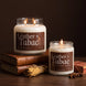 Leather + Tabac 18 oz and 8 oz soy candles by Upsensed, lit, the 18 oz on stacked leather-bound books, with dried tobacco leaves, a cherrywood block, and star anise on a walnut table against a dark espresso backdrop.