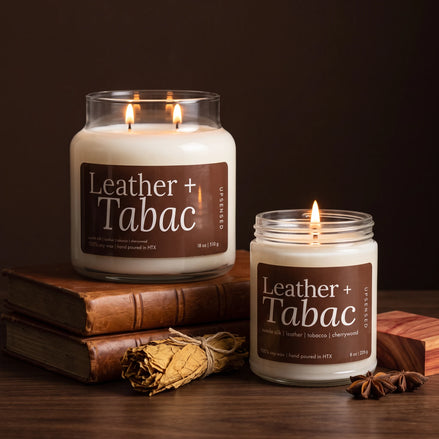 Leather + Tabac 18 oz and 8 oz soy candles by Upsensed, lit, the 18 oz on stacked leather-bound books, with dried tobacco leaves, a cherrywood block, and star anise on a walnut table against a dark espresso backdrop.