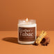 Leather + Tabac 8 oz soy candle by Upsensed, lit, in a clear glass jar with a brown label listing suede silk, leather, tobacco, and cherrywood notes, beside a bundle of dried tobacco leaves, a cherrywood block, and star anise on a warm cognac backdrop.