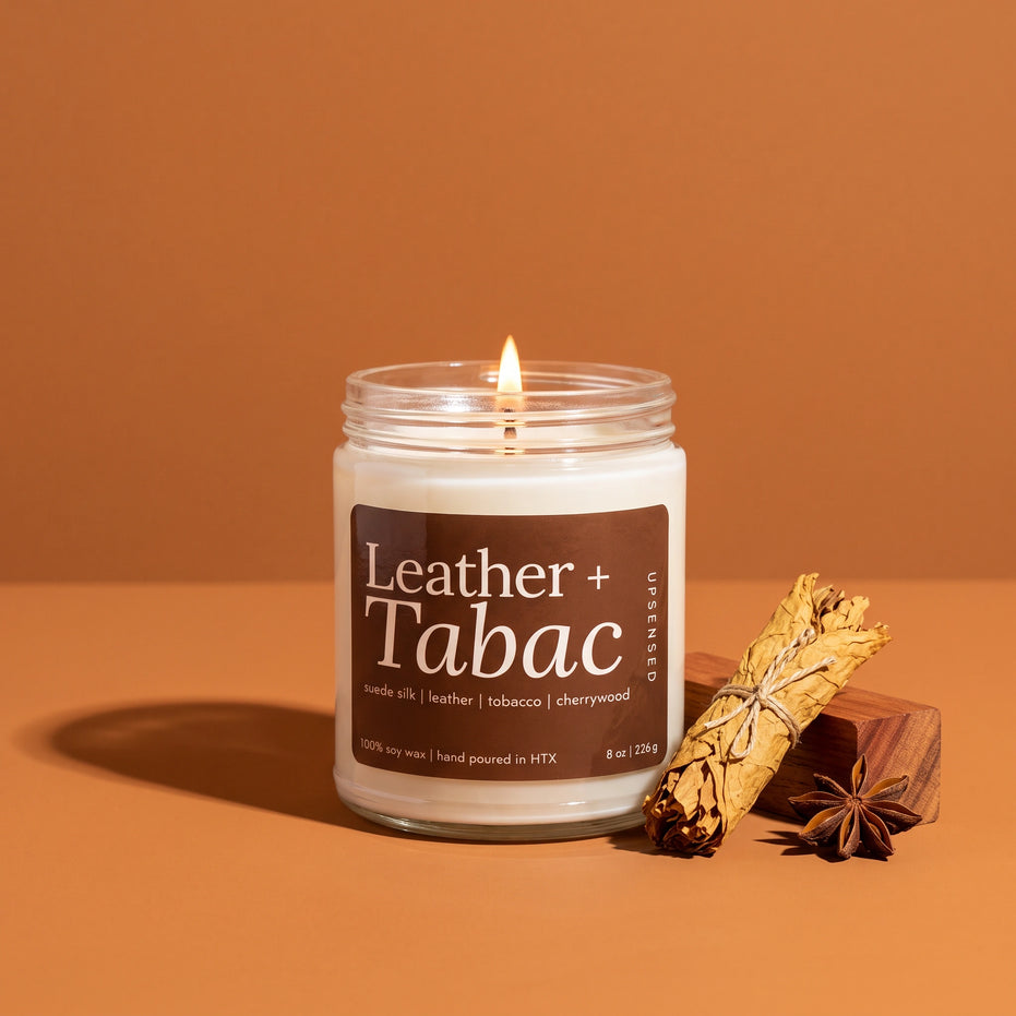 Leather + Tabac 8 oz soy candle by Upsensed, lit, in a clear glass jar with a brown label listing suede silk, leather, tobacco, and cherrywood notes, beside a bundle of dried tobacco leaves, a cherrywood block, and star anise on a warm cognac backdrop.