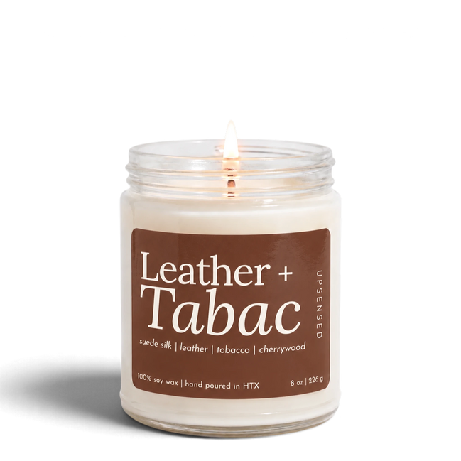 Leather + Tabac 8 oz soy candle by Upsensed, lit, in a clear glass jar with a brown label listing suede silk, leather, tobacco, and cherrywood notes.