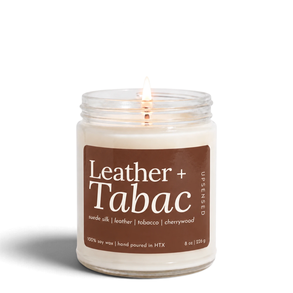 Leather + Tabac 8 oz soy candle by Upsensed, lit, in a clear glass jar with a brown label listing suede silk, leather, tobacco, and cherrywood notes.