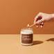 A hand with gold rings and caramel nails lighting a Leather + Tabac 8 oz soy candle by Upsensed with a wooden match, on a two-tone cognac and sand set with soft sunlight.