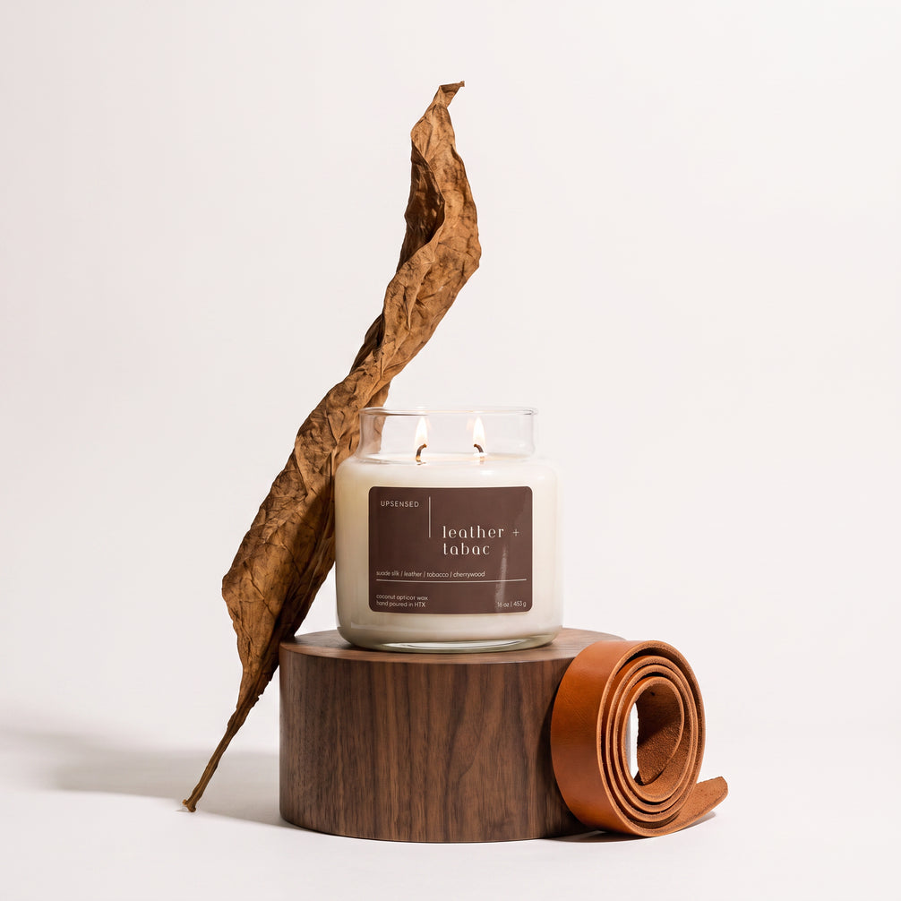Upsensed Leather + Tabac candle burning on a round wooden pedestal, styled with a curled dried tobacco leaf and a coiled leather strap.
