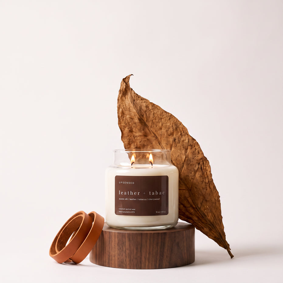 Upsensed Leather + Tabac candle burning on a round wood pedestal, styled with a large dried tobacco leaf and a coiled leather strip.