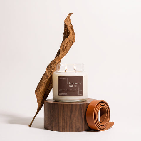 Upsensed Leather + Tabac candle burning on a round wooden pedestal, styled with a curled dried tobacco leaf and a coiled leather strap.