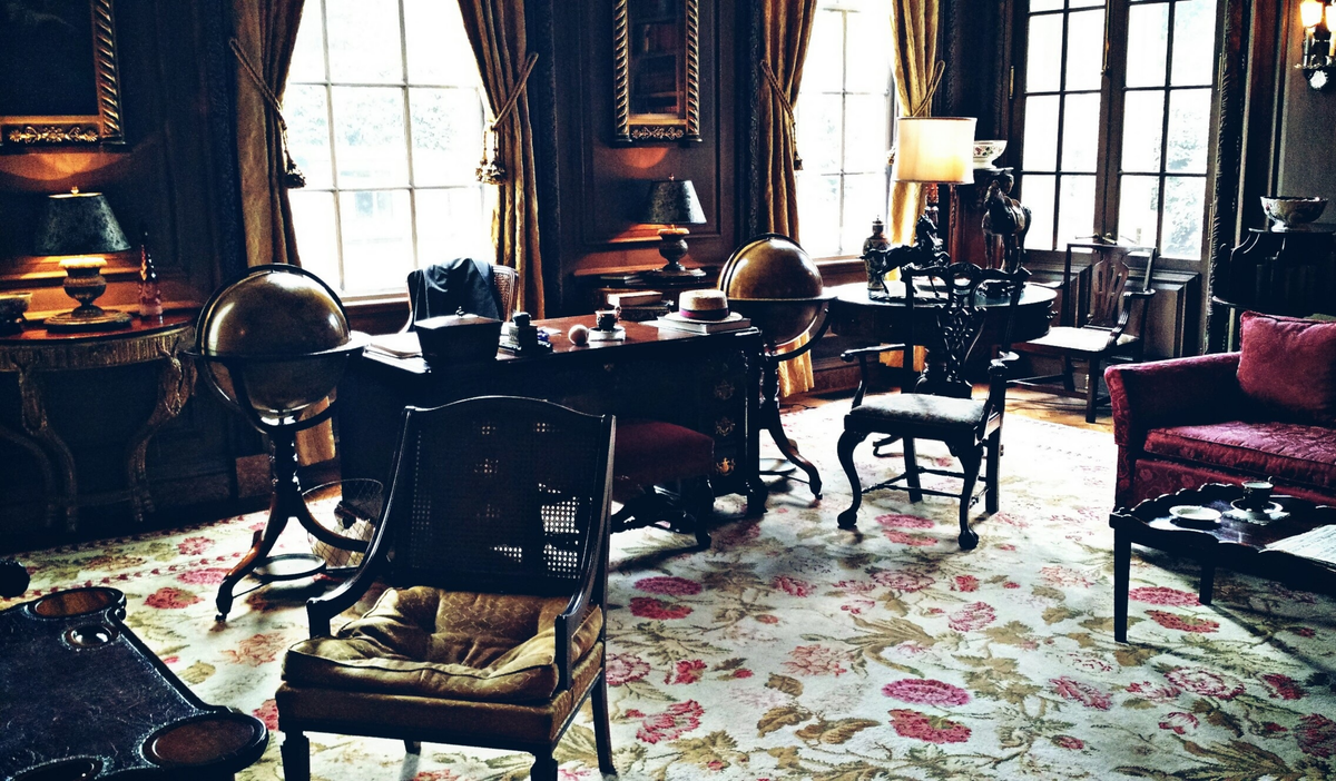 An ornate vintage study or library interior with dark wood desks, chairs, lamps, large windows with drapes, a floral patterned rug, and a red upholstered armchair.
