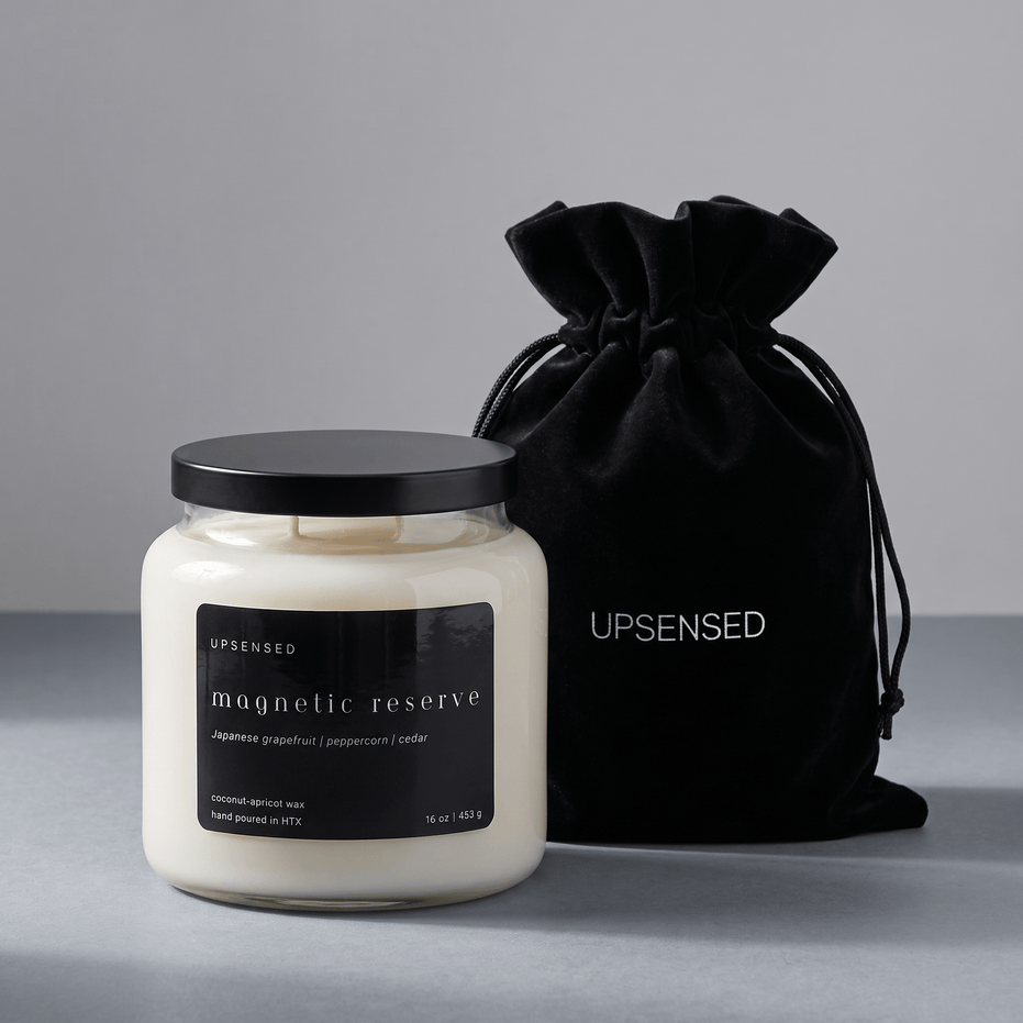 Upsensed Magnetic Reserve candle with a black lid beside its black velvet UPSENSED drawstring packaging bag, on a grey background.