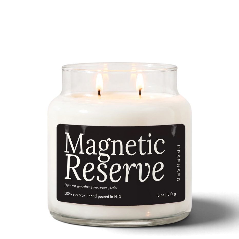 Magnetic Reserve 18 oz double-wick soy candle by Upsensed, both wicks lit, in a clear glass jar with a black label listing Japanese grapefruit, peppercorn, and cedar notes.