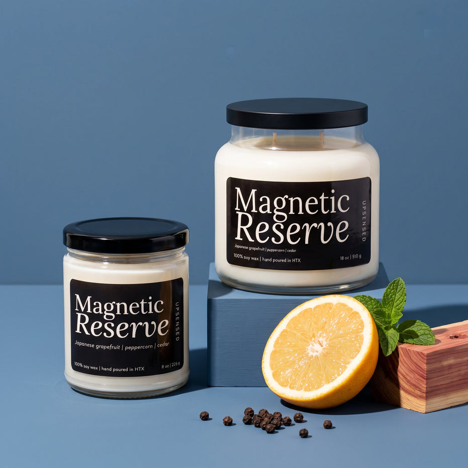 Magnetic Reserve soy candles by Upsensed in 8 oz and 18 oz sizes with black lids and black labels, the 18 oz raised on a blue block, styled with a halved grapefruit, fresh mint, cedar wood, and black peppercorns on a slate blue backdrop.