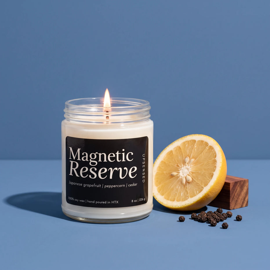 Magnetic Reserve 8 oz soy candle by Upsensed, lit, in a clear glass jar with a black label listing Japanese grapefruit, peppercorn, and cedar notes, beside a halved grapefruit, a wood block, and black peppercorns on a blue backdrop.