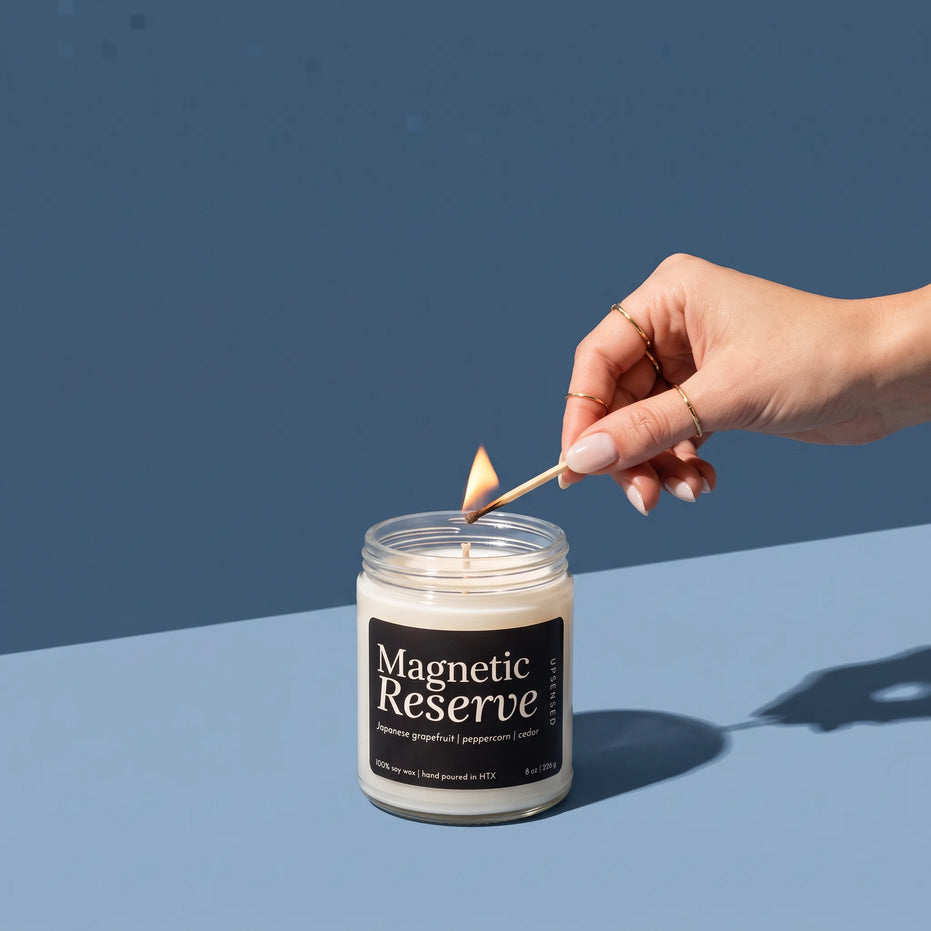 A hand with gold rings lighting a Magnetic Reserve 8 oz soy candle by Upsensed with a wooden match, on a two-tone blue set with hard sunlight and long shadows.