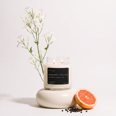 Upsensed Magnetic Reserve candle burning on a round ceramic base, styled with jasmine flower stems, black peppercorns, and a halved grapefruit.
