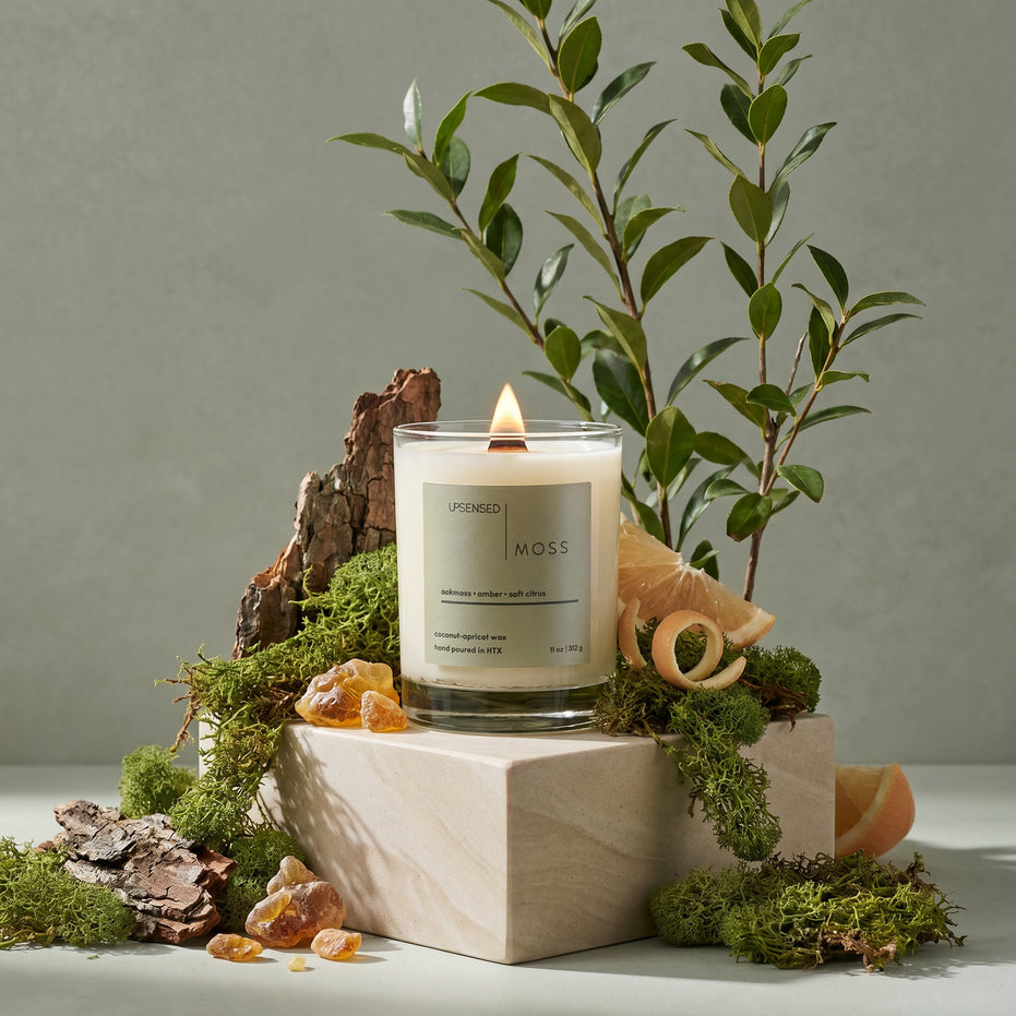 Upsensed Moss signature candle burning on a stone pedestal surrounded by oakmoss, amber resin, citrus slices, and green botanicals; scented with oakmoss, amber, and soft citrus in coconut-apricot wax, hand poured in Houston