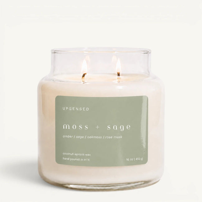 Upsensed Moss + Sage 16 oz double-wick candle in a clear apothecary jar with a sage green label, amber, sage, oakmoss, and rose musk scent notes, on a plain white background.