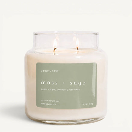Upsensed Moss + Sage 16 oz double-wick candle in a clear apothecary jar with a sage green label, amber, sage, oakmoss, and rose musk scent notes, on a plain white background.