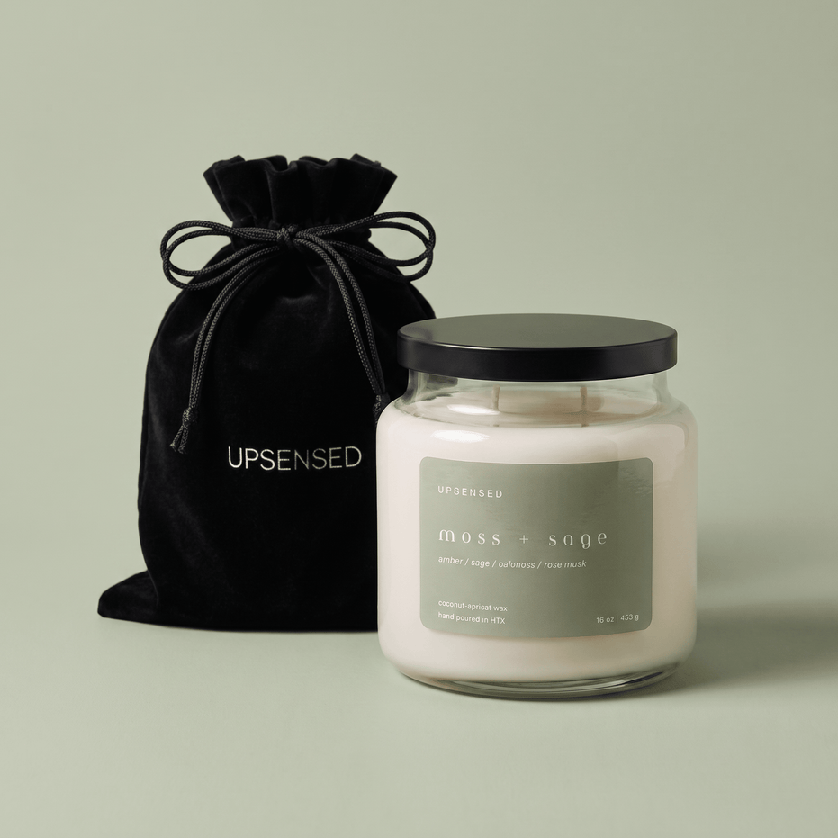 Upsensed Moss + Sage candle with a black lid beside its black velvet UPSENSED drawstring packaging bag, on a sage green background.