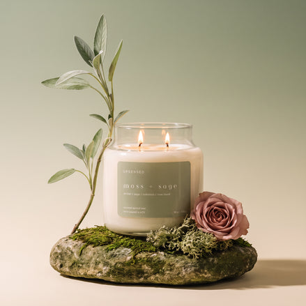 Upsensed Moss + Sage candle burning on a moss-covered stone, styled with a sage sprig and a dusty pink rose against a green gradient background.